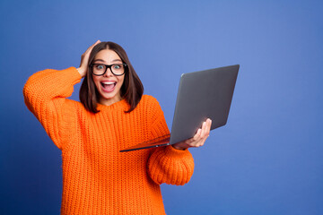 Excited young woman holding laptop in orange sweater against purple backdrop, expressing surprise and joy in casual style.