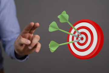 Image of a dartboard with three darts hitting the bullseye and a hand pointing towards the target,...