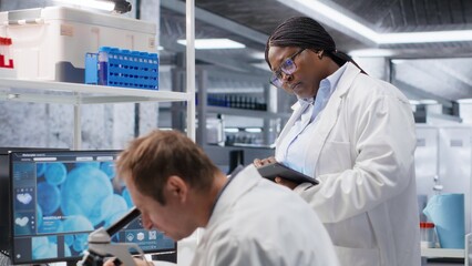 Employees team studying sample tray under the microscope for research study in a lab, cellular...