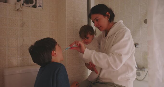 Mother holding baby while teaching son oral hygiene brushing teeth in bathroom, daily parenting care and multitasking family routine at home