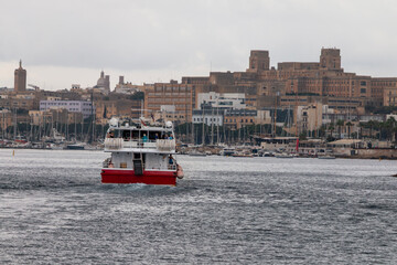 Fototapeta premium Malta - A red and white ferry boat transports passengers across a harbor, with a historic city skyline, numerous buildings, and sailboats visible under an overcast sky.