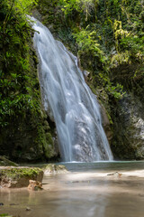 A serene waterfall cascades over a mossy rock face into a calm pool amidst a lush green forest.