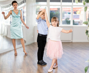 Naklejka premium Couple of positive interested preteen dancers, boy and girl preparing for performance, rehearsing ballroom dance in spacious classroom while female teacher observing from background