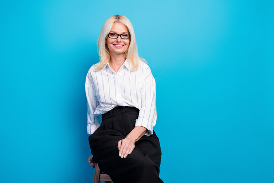 Professional mature businesswoman smiles confidently in a striped blouse and black skirt posing on a stool for corporate office lifestyle image