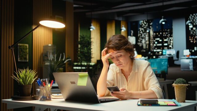 Weary businesswoman stays connected on social media while she does overtime at night, checking her mobile phone after receiving a notification. Female worker checks text messages. Camera A.