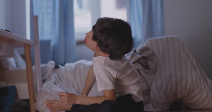 Boy sitting on bedroom floor looking up with expression of boredom, authentic childhood moment of restlessness, real life emotion and everyday family home environment