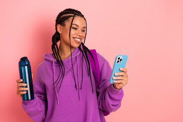 Young woman with braids holds a water bottle and checks her phone against a pink background,...