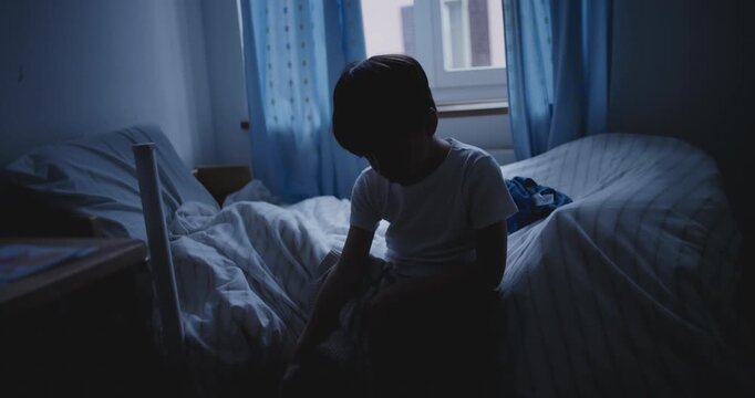 Child sitting alone in dark bedroom with head bowed, symbolizing sadness, loneliness, and childhood depression
