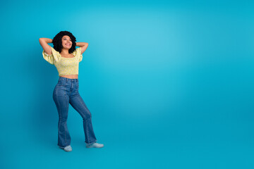 Young woman posing with hands on head against a blue backdrop, wearing a yellow top and jeans