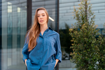 Young woman with long hair in blue shirt standing with hands in pockets looking at camera in front of modern office building. She exudes confident and smart casual style in corporate setting