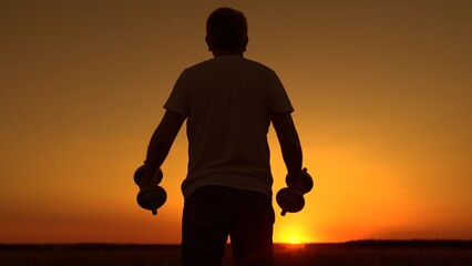 Person exercises for healthy body. Man exercises with dumbbells in park at sunset. Healthy adult man exercises outdoors. Man does strength training, dumbbell deadlifts, bicep curls. Nature Fitness.