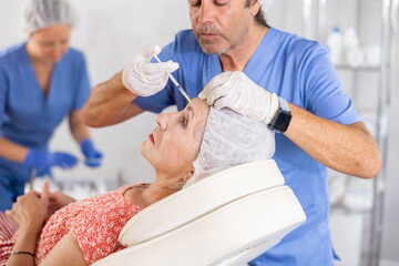 Senior woman undergoing botulinum therapy session at aesthetic medicine clinic. Skilled male cosmetologist making injections between eyebrows of patient with insulin syringe to correct facial wrinkles