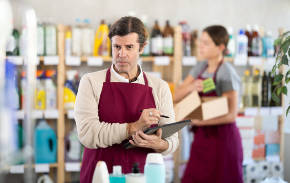 Focused supermarket manager in burgundy apron conducting inventory using tablet to check of cleaning and personal care products, with female employee organizing items in background - Powered by Adobe