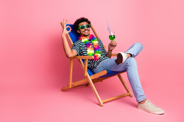 Young mixed race man relaxing in a colorful deck chair with lei sunglass and green drink on pink studio background