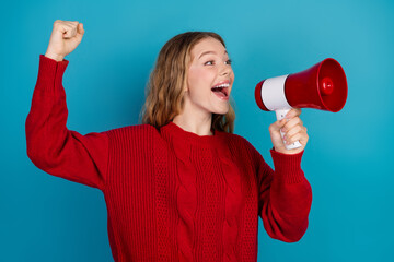 Adorable young woman in a vibrant red sweater enthusiastically holding a megaphone against a blue...