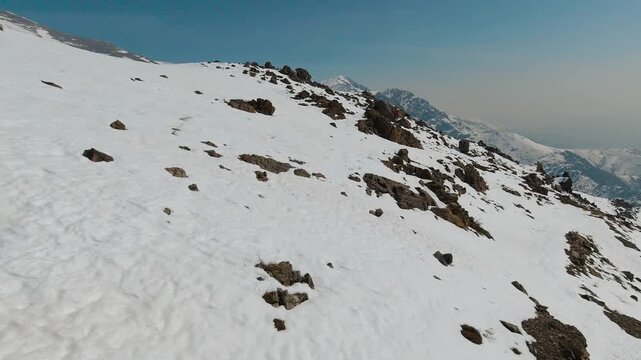Aerial tracking shot camera moves up above the ridge of a high mountain in late winter. Patchy melting snow reveals brown rock and a thin winding path across the slope, suggesting isolation, endurance