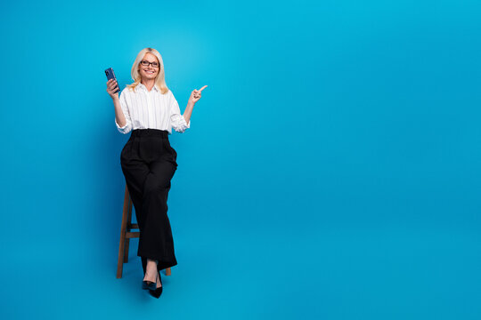 Businesswoman in white shirt and black pants sits on a stool against a blue background holds a smartphone and smiles while pointing