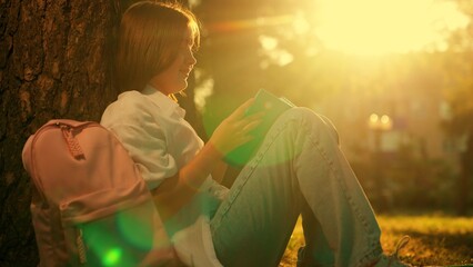 Schoolgirl sits with book under tree on park lawn. Education. Teenage schoolgirl with school backpack reads textbook while sitting in city park. Child relaxes with book on in nature park. Education