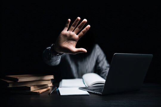 Person making a stop gesture, preventing access or seeking privacy while working in a dark environment with a laptop and books