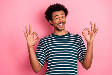 Young mixed race man winking and smiling in striped shirt against pink background showcasing casual...