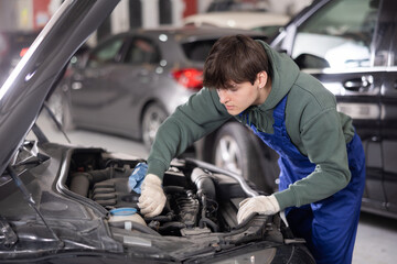 Young male auto mechanic in blue overalls and gloves focused on adjusting parts and tightening...