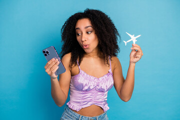Young woman in purple top holds smartphone and model airplane against blue background conveying...