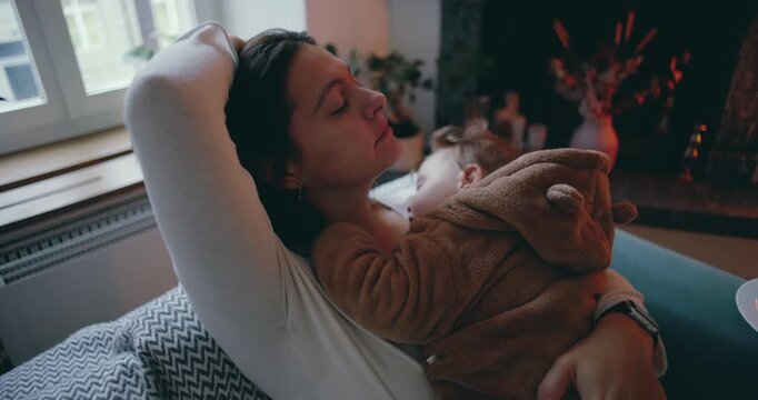 Mother resting on couch with sleeping baby in arms, tired but serene, symbol of unconditional maternal love, peaceful domestic moment of comfort and emotional renewal