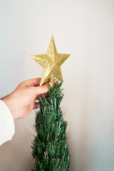 Hand of an unrecognizable person placing a bright golden glitter star on top of the plastic Christmas tree