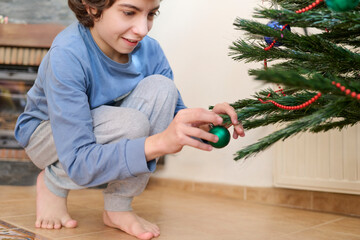 Child decorates the Christmas tree with red and shiny ornaments with excitement while wearing his pajamas