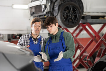 Serious looking middle aged man, car service owner in blue overalls and plaid shirt with pneumatic wrench in hands giving vehicle repair tips to young puzzled mechanic