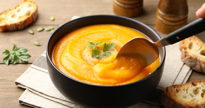 Woman taking tasty pumpkin cream soup with spoon from bowl at wooden table, closeup