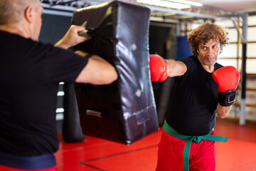 Green-belt Krav Maga student, dressed in red training pants, black T-shirt and boxing gloves, driving straight punch into partners padded shield during focused self-defence drill in gym