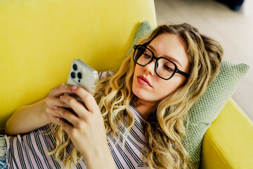 Relaxed woman with glasses lounges on a bright yellow couch at home using a smartphone in a cozy...