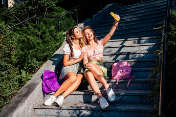 Two young friends take a sunny selfie on city stairs while traveling together enjoying fashion and summer vibes
