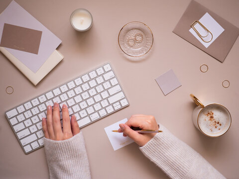 A feminine workspace flat lay top view on a calm beige background, featuring a close-up of a woman's hands typing on a wireless keyboard while taking notes.