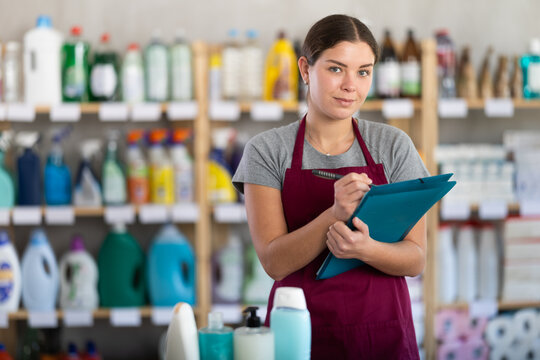 Thoughtful young female store supervisor in burgundy apron taking notes on clipboard while taking inventory of cleaning and hygiene products
