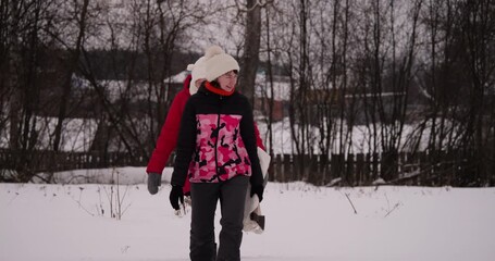 Two girls carrying axes and white bag walking through deep winter snow towards forest. Village in background Christmas tree tradition celebration - Powered by Adobe