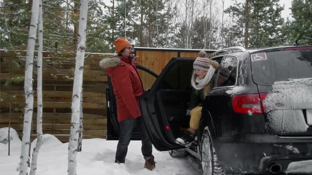 Full shot of young Caucasian father with missing arm disability and son in warm jackets and hats arriving for winter vacation at forest cabin, getting out of car, looking around at snow in excitement