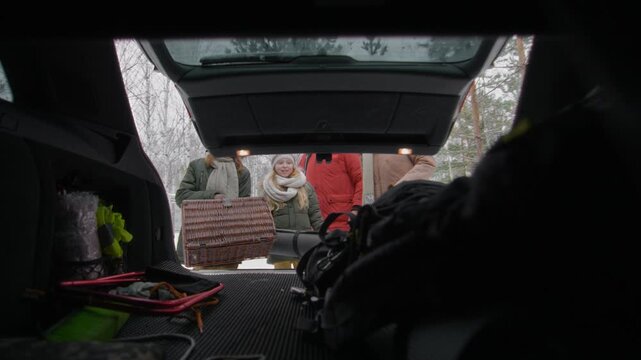 Medium full shot of young Caucasian family loading luggage bags, backpack and red sled into hatchback car trunk, while departing to go home after winter holiday at forest cabin