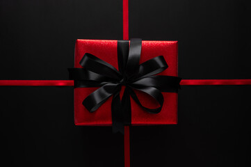 Black Friday with a stunning red gift box with a black satin bow is centered on crossing red ribbons on a dark black background.