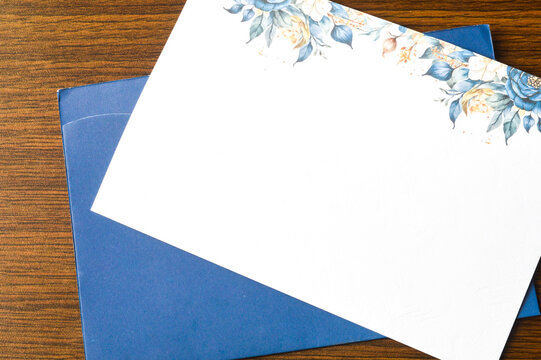 Blank white greeting card or wedding invitation with watercolor blue and gold floral borders, resting on a dark blue envelope and wooden background. Ideal mockup for events and stationery.