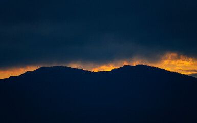 A dramatic sunset with a sliver of bright orange light on the horizon below a dark mountain silhouette and heavy storm clouds