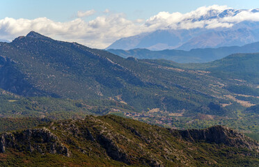 A scenic view of a mountain range with peaks touching the soft clouds and a lush green forested slope in the foreground