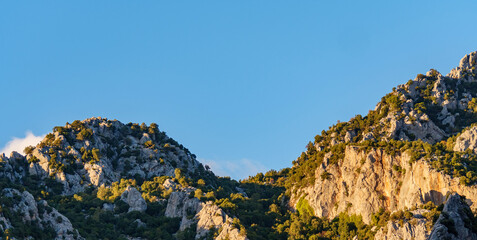 A panoramic view of a beautiful mountain range with rocky peaks and green trees under a blue sky with soft white clouds