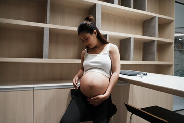 An eight-month pregnant Asian woman sits rubbing her belly in the kitchen of a condominium.
