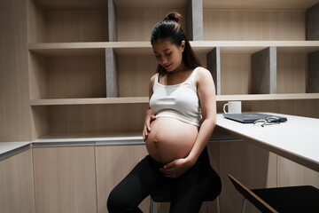 An eight-month pregnant Asian woman sits rubbing her belly in the kitchen of a condominium.
