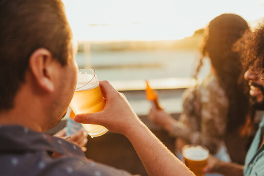 Close-up of a man raising a glass of beer as he enjoys a relaxed sunset moment on a rooftop with friends.