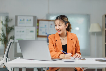 Asian businesswoman on a bright office video call, smiling and taking notes at her laptop desk during a virtual meeting
