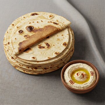 A stack of thin, pliable laffa bread (Middle Eastern flatbread) fresh off the saj oven, with a slight char, next to a bowl of hummus. Soft, textured fabric underneath