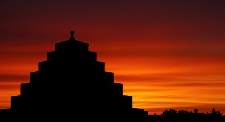 An iconic tiered architectural structure stands dramatically against the fiery orange and red evening sky, creating a striking black silhouette ,majestic ,asian ,landscape